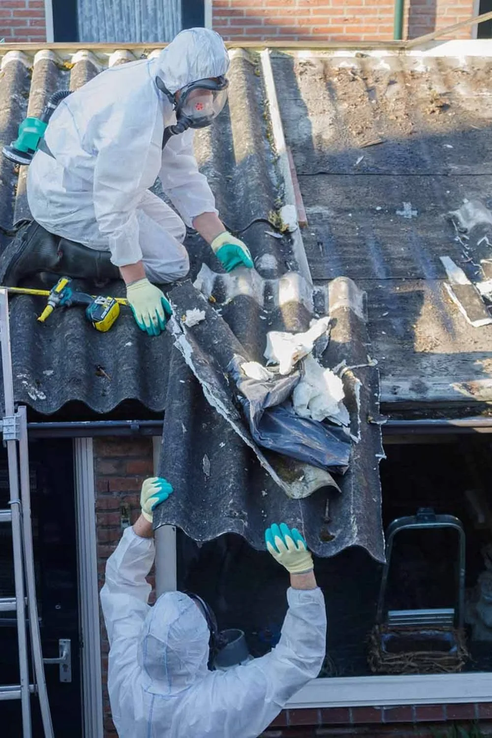 Two workers in protective suits lowering a whole asbestos roof sheet by hand