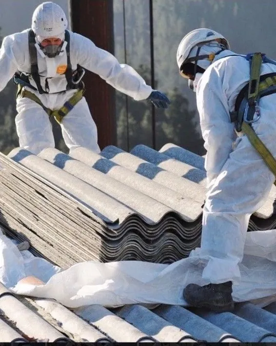 Two workers in white protective suits, respirators and harnesses wrapping corrugated roof sheets in plastic on a roof